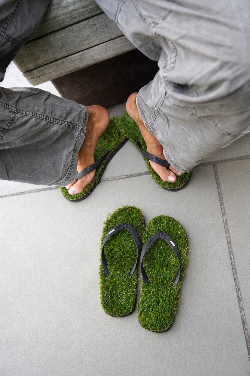 A pair of moss green flip-flops with black straps, shown on a tiled floor with a person's legs crossed over them wearing matching moss green flip-flops.